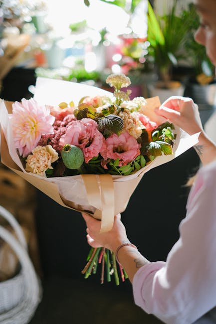 A person is holding a large, colorful floral bouquet featuring pink and peach peonies, cream carnations, white and purple statice, and green foliage arranged in a loosely styled bouquet. The flowers a