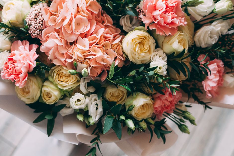 A floral arrangement featuring a variety of fresh flowers, including soft pink hydrangeas, cream-colored roses, and white lisianthus, all elegantly arranged with dark green foliage. The bouquet is wra