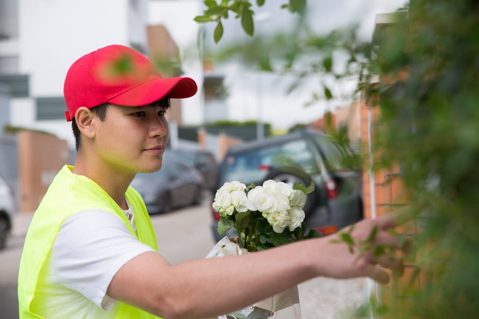A young male flower delivery person wearing a red cap and a neon yellow vest is arranging a bouquet of fresh white hydrangeas wrapped in brown paper. He is standing outdoors on a residential street, w