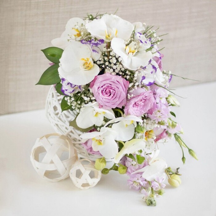 A lavish bridal bouquet in a cascading, teardrop shape is presented against a light, neutral backdrop, its form flowing downward like a gentle waterfall of petals and foliage. At the top, the flowers gather densely around what appears to be a white decorative base or small pedestal with an intricate, lace-like or embossed pattern, giving the impression of delicate fabric or finely crafted ceramic. From this anchor point, large white orchids resembling Phalaenopsis arc outward and downward, their petals broad, smooth, and luminous, with touches of soft yellow and gold in their centres adding a sophisticated, exotic accent. Nestled among the orchids are roses in tender shades of blush pink, pale lavender, and soft lilac, each bloom layered with velvety petals at varying stages of opening, creating a rich, romantic texture. Fine sprigs of white baby's breath weave through the entire arrangement, softening transitions and giving the bouquet a dreamy, slightly airy appearance. Additional blossoms in cool lilac and light purple tones, likely lisianthus or similar ruffled flowers, tuck in around the roses and orchids, enhancing the pastel colour story and adding intricate detail. As the bouquet tapers downward, blooms and foliage gradually become sparser, forming an elegant point that suggests natural movement. Touches of fresh green leaves appear here and there, grounding the pastels and adding freshness without overwhelming the design. Around the base on the pale surface rest several white ornamental spheres with ornate cut-out patterns, like sculpted lace lanterns, reinforcing the refined, bridal mood. The soft cream background remains intentionally unobtrusive, slightly out of focus, so all attention settles on the shapes, colours, and craftsmanship. This is the kind of cascading bouquet that would suit a stylish Barnsbury wedding reception or a ceremony near Islington's leafy squares, capturing both classic romance and modern sophistication.